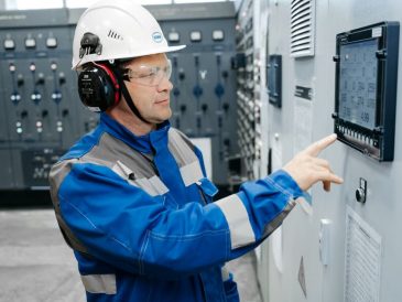 Technician in safety gear working at an industrial control panel indoors.